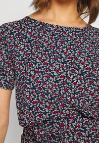 Marinblus med korta ärmar, prydd med röda och krämfärgade blommor på en mörk bakgrund. Mjuk tyg, lös passform, rund halsringning.