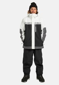 Giacca da neve in bianco e nero con dettagli grigi, dotata di colletto alto, tasche con zip e pantaloni svasati. Tessuto testurizzato, resistente alle intemperie.