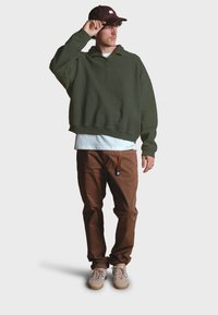 Sudadera verde de media cremallera, camiseta blanca, pantalones marrones y zapatos beige. La gorra tiene un logo y los pantalones cuentan con una etiqueta lateral. Corte relajado.