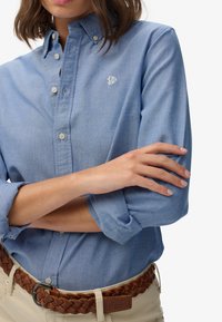 Jasnoniebieska koszula z kołnierzykiem, z długim rękawem podwiniętym, oraz małym haftowanym logo. Nosi ją z plecionym brązowym paskiem na beżowych spodniach.