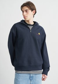 Μπλε ναυτικό πουλόβερ με φερμουάρ και όρθιο λαιμόκοψη, με λογότυπο μπροστά και μανίκια με μανσέτες, φορεμένο πάνω από ριγέ πουκάμισο και ανοιχτόχρωμο τζιν.