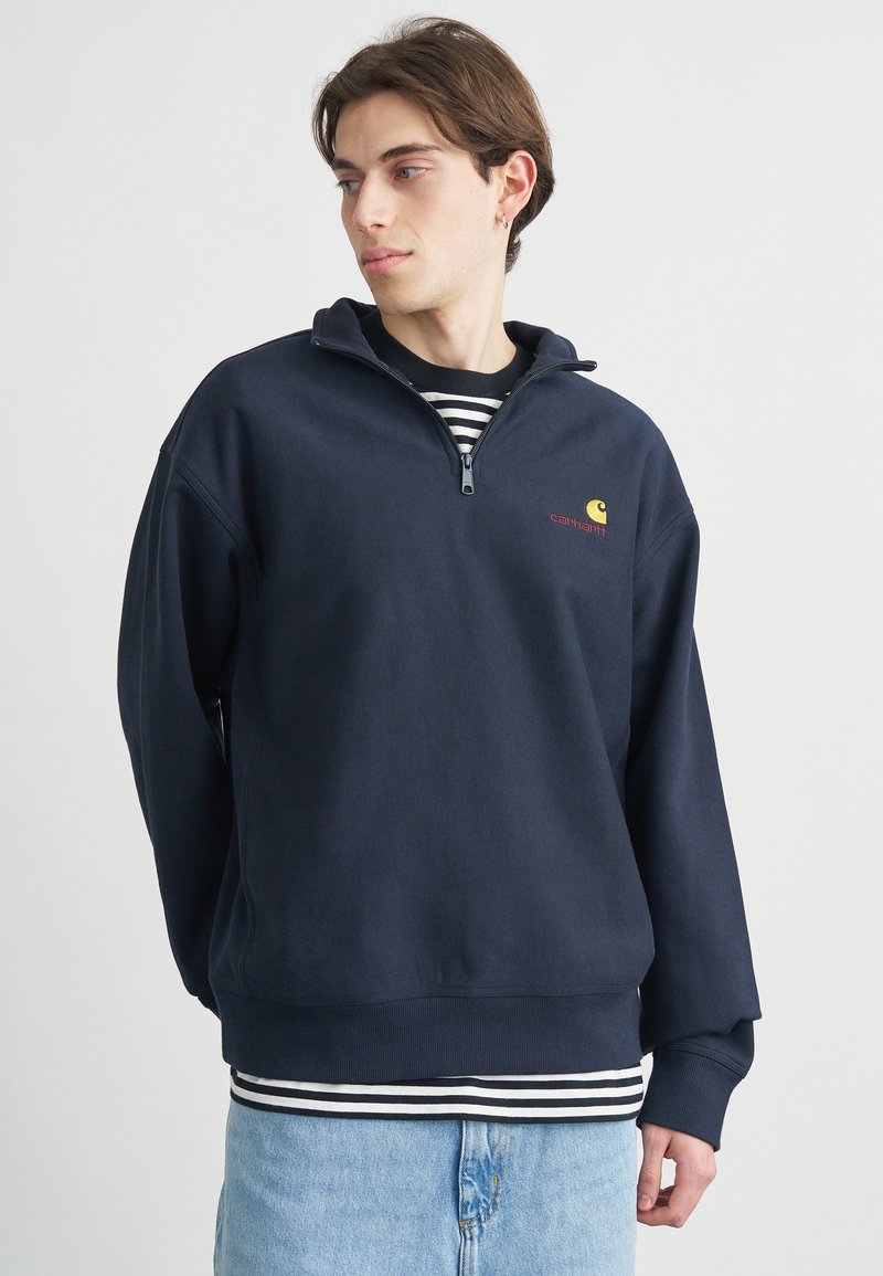 Μπλε ναυτικό πουλόβερ με φερμουάρ και όρθιο λαιμόκοψη, με λογότυπο μπροστά και μανίκια με μανσέτες, φορεμένο πάνω από ριγέ πουκάμισο και ανοιχτόχρωμο τζιν.