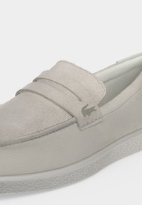 Mocassin en daim beige clair avec une bride sur le dessus et un petit logo crocodile sur le côté près du talon.
