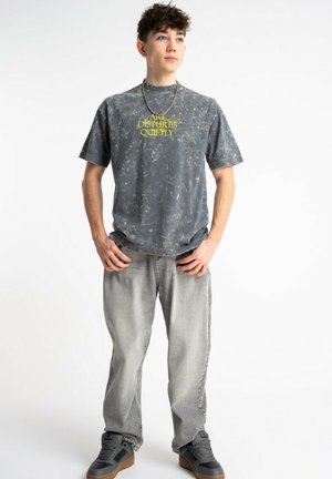 Młody mężczyzna stoi z rękami w kieszeniach, ubrany w wyblakły czarny t-shirt z grafiką, jasnoszare jeansy, naszyjnik z łańcuszka i szare tenisówki.