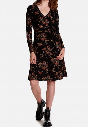 Femme portant une robe portefeuille noire à fleurs avec des manches longues et des bottines noires, debout avec une jambe légèrement en avant.