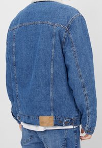 Muž v modré džínsové bundě s ovčími límcem a odpovídajícími modrými džíny, pohled zezadu proti jednobarevnému pozadí.