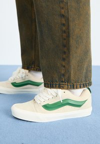 Beige suede sneakers with green leather side stripe, white rubber sole, and lace-up style. Worn with dark denim pants against a blue floor.