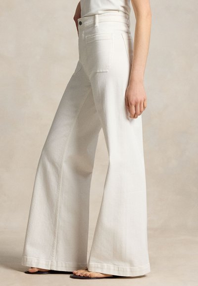 Γυναίκα που φοράει λευκά τζιν με ψηλόμεση και φαρδιά μπατζάκια και μπεζ σανδάλια, στέκεται πλάγια μπροστά σε ουδέτερο φόντο.