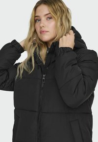 Veste rembourrée noire avec capuche, dotée d'une fermeture éclair à l'avant, de poignets élastiques et de poches latérales. Tissu lisse avec une finition mate.