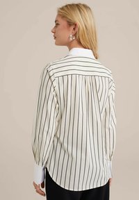 Camicia bianca a maniche lunghe con strisce verticali nere, presenta un colletto a punta e polsini abbottonati, e un design con pieghe sul retro.