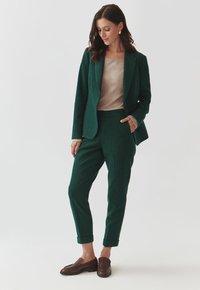 Green wool blazer and matching tapered pants, paired with a beige top. Brown loafers complete the outfit. Textured fabric and rolled cuffs visible.