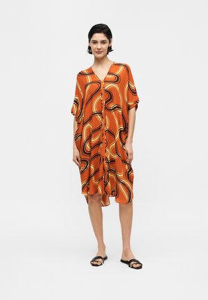 Nő áll, narancssárga térdig érő ruhában, fekete és sárga absztrakt spirálokkal, fekete nyitott orrú szandálban, semleges háttér.