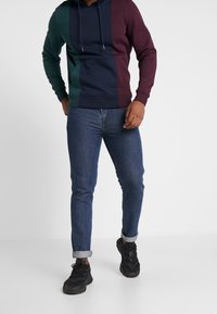 Sudadera a bloques de color en verde oscuro, azul marino y burdeos, con un bolsillo tipo canguro en el frente y capucha con cordón, combinada con jeans de denim enrollados y zapatillas negras.