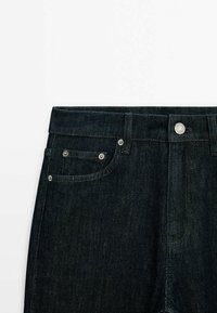 Jean en denim bleu foncé avec fermeture à boutons, poches avant visibles et passants de ceinture sur fond blanc.