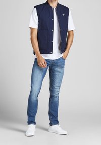 Navy blue sleeveless vest with snap buttons, paired with a white T-shirt, light blue jeans, and white sneakers. Smooth fabric and casual fit.