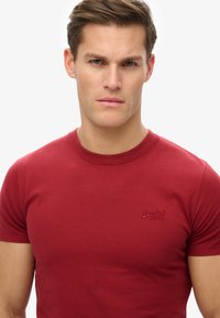 Camiseta roja de manga corta fabricada en algodón, con un cuello redondo y un pequeño logotipo bordado en el pecho. Textura suave, diseño ajustado.