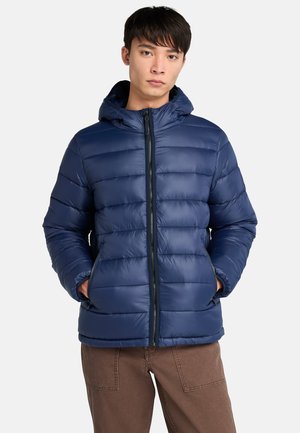 Giovane uomo in un giubbotto a vento blu navy con cappuccio, in piedi con le mani in tasca, su uno sfondo bianco uniforme.