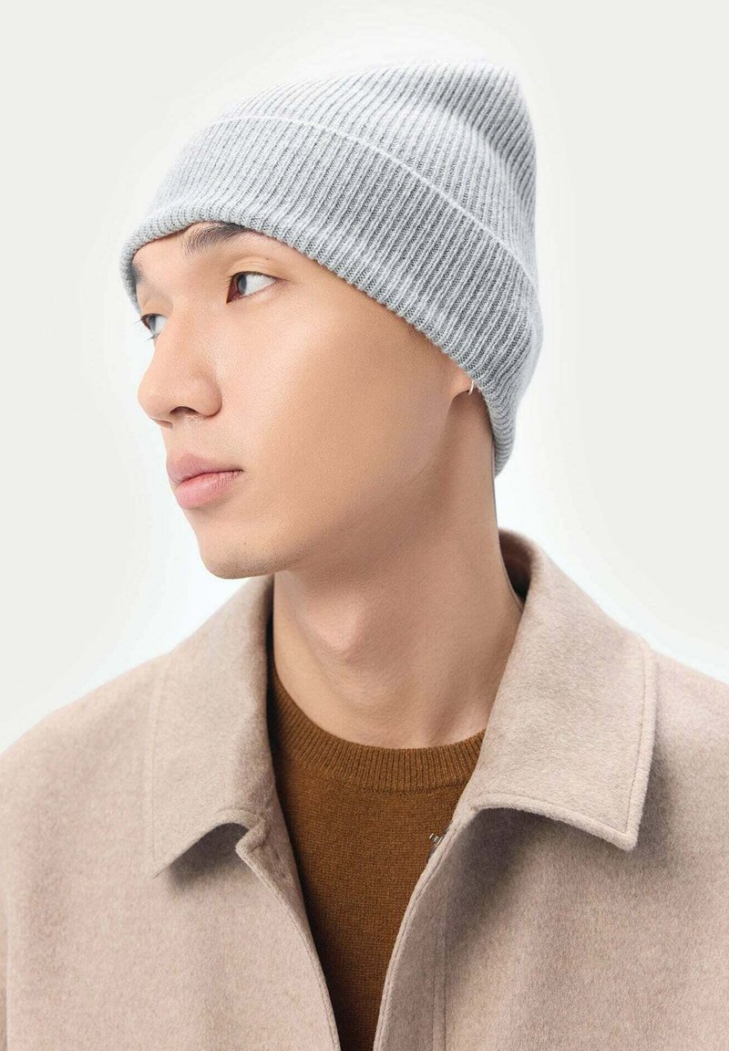 Grå ribbad stickad mössa med en avsmalnande design. Bar över en ljusfärgad kappa ovanpå en brun tröja. Slät textur, minimala accenter.