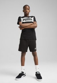 Černé bavlněné tričko s bílým logem Puma, doplněné černými šortkami. Celkový vzhled doplňují černé tenisky, bílé ponožky a neutrální pozadí.