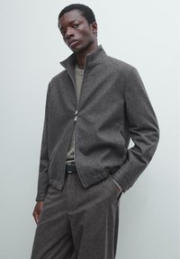 Graue Reißverschlussjacke mit hohem Kragen, aus strukturiertem Stoff und elastischen Bündchen, kombiniert mit einem grauen Pullover und einer Hose.