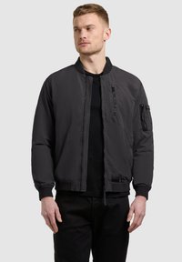 Homme portant une veste bomber noire avec plusieurs poches zippées, debout les mains détendues, regardant légèrement sur le côté sur un fond uni.