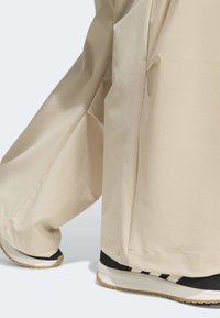 Béžové kalhoty s širokými nohavicemi z měkké látky, s hlubokými bočními kapsami, doplněné černo-bílými teniskami s texturovanými detaily.