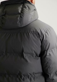 Donkergrijze pufferjack met een grote capuchon, voorzien van gestikte secties en een subtiele logo-detail op de schouder. Zachte, matte textuur.