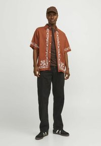 Chemise brune à manches courtes avec broderies florales blanches, portée sur une chemise marron foncé, associée à un jean noir et des baskets noires.