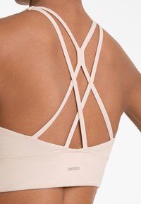 Beige sportbh met gekruiste rugbanden, een gladde textuur, zachte stof en een klein logo op de band.