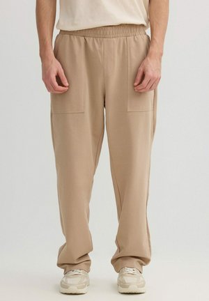 Pantalones casuales beige con cintura elástica y bolsillos delanteros de parche, combinados con zapatillas beige y una camisa clara en una persona de pie.