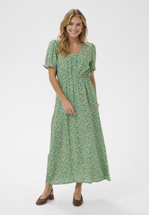 Uśmiechnięta kobieta w zielonej, kwiatowej sukience maxi z krótkimi bufkami i brązowych plecionych butach na obcasie, stojąca na tle białego tła.
