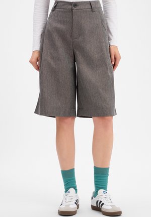 Frau trägt graue knielange Shorts, türkisfarbene Socken, weiße Turnschuhe mit schwarzen Streifen und ein weißes Langarmshirt, steht vor einem einfarbigen Hintergrund.
