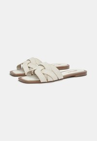 Sandales à glissière beige clair avec un design de sangle tressée, semelle plate et texture douce. Bout arrondi et subtils accents marron sur la semelle.