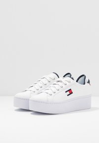 Baskets plates blanches avec une texture en cuir lisse, un design classique à lacets et un petit logo avec des accents rouges, blancs et bleus.