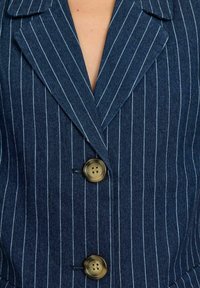 Tumedast sinise pinstripe'iga bleiser, millel on nurgeline kaelus, kaks suurt pruuni nööpiga ja tekstuuriga kangas.