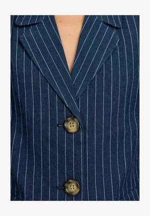 Mornarsko modra blazerja z navpičnimi črtami z izrezanim ovratnikom, ki ima dva velika rjava gumba in teksturirano tkanino.