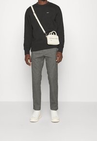Homme portant un sweat-shirt noir, un pantalon gris, des baskets blanches et un petit sac bandoulière blanc sur un fond blanc uni.
