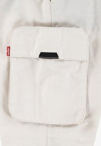 Beige tyg med en stor framficka som har en svart accent. En röd logotagg syns på vänster sida av fickan. Slät textur.