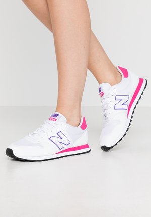 Person iført hvide New Balance 500 sneakers med lyserøde og lilla detaljer, stående med den ene fod let løftet på en ensartet overflade.