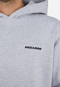 Sudadera gris de tejido suave con bolsillo canguro. Presenta un logotipo negro "PEGADOR" bordado en el lado izquierdo del pecho.
