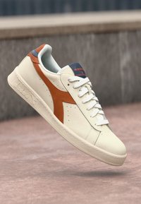 White leather sneaker with an orange and navy accent. Features a rounded toe, flat laces, and a textured sole. Simple design and stitching detail.