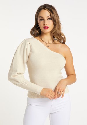 Giovane donna con capelli ondulati fino alle spalle che indossa un maglione crema monospalla con manica a sbuffo e pantaloni bianchi.