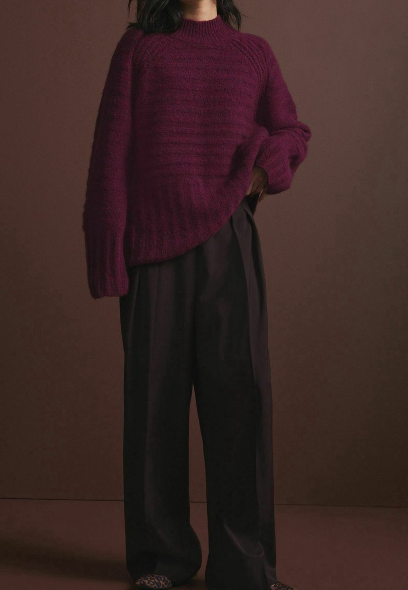 Personne portant un pull en maille bordeaux oversize et un pantalon noir ample, debout devant un fond marron uni.