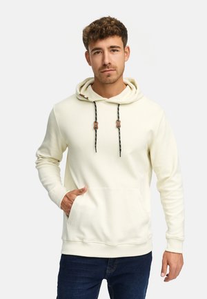 Crème hoodie met ribbeltextuur, verstelbare capuchon met zwarte trekkoorden, voorste kangoeroezak en ribbelmanchetten. Gecombineerd met donkere jeans.
