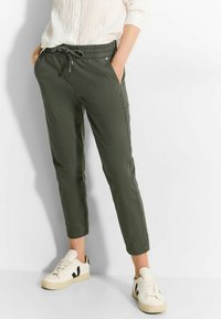 Olijfgroene, taps toelopende joggingsbroek met een elastische tailleband, trekkoord en zijzakken; gecombineerd met witte sneakers met zwarte accenten.