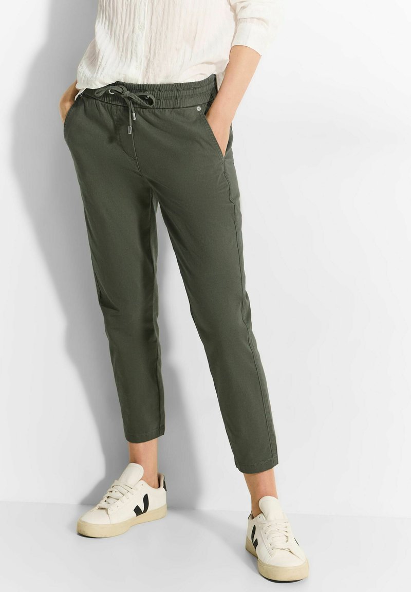 Olijfgroene, taps toelopende joggingsbroek met een elastische tailleband, trekkoord en zijzakken; gecombineerd met witte sneakers met zwarte accenten.