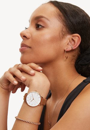 Brauner Lederarmbanduhr mit weißem Zifferblatt und roségoldenen Akzenten. Das Modell trägt kleine Creolen und ein zartes Kettenarmband.