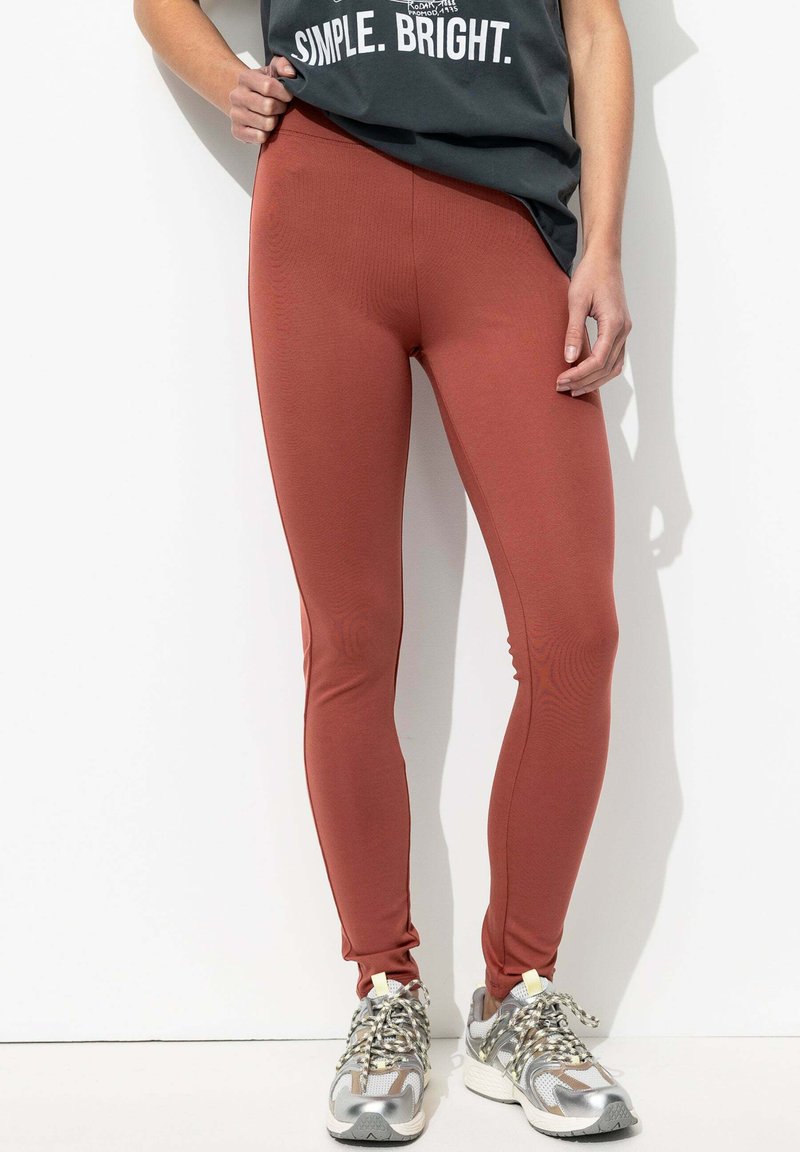 Leggings rouge rouille en tissu extensible, avec une texture lisse et un design ajusté, associés à des baskets grises et argentées.
