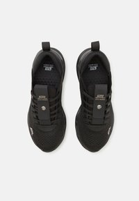 Paire de baskets de sport noires Steve Madden avec empeignes en maille, sangles élastiques à boutons-pression, et détails du logo sur la languette et les orteils.