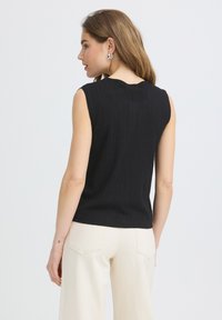 Femme aux longs cheveux portant un débardeur noir côtelé sans manches et un pantalon écru, vue de dos sur un fond clair uni.